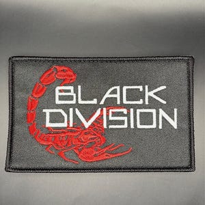 Op de afbeelding: Een rechthoekige zwarte geborduurde patch met een rode schorpioenafbeelding aan de linkerkant en de witte tekst "BLACK DIVISION" in een gestileerd lettertype in het midden, afgewerkt met een zwarte gestikte rand rondom het geheel.