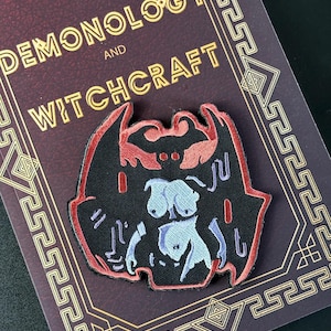 Può includere: Una toppa nera con bordo rosso e un'illustrazione stilizzata di un demone. Il demone ha la pelle azzurra e dettagli rossi. La toppa è su un libro con le parole "Demonology and Witchcraft" in lettere dorate.