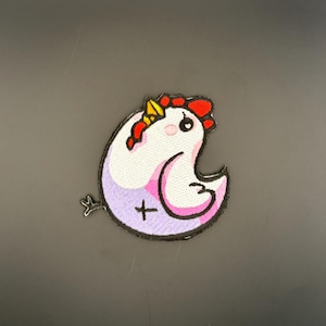 Op de afbeelding: Geborduurde patch met een grillig kippenontwerp. De patch heeft een witte body met roze en paarse accenten, een rode kam en een gele snavel. Een zwarte omtrek definieert de vorm en een klein kruis is zichtbaar.