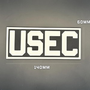 May include: A rectangular patch featuring the text USEC in bold white block letters on a black background. The patch has a white border and measures 140mm in width by 60mm in height as indicated by the text labels on the image.