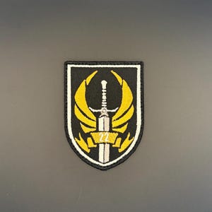Op de afbeelding: Geborduurde patch met een zwarte schildvorm, met een zwaard en gele vleugels. De patch bevat het nummer "22" in het midden. De rand is wit en de achtergrond is zwart.