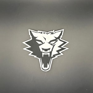 May include: A black and white sticker depicting a snarling wolf's head. The wolf's face is black with white highlights and a white outline. The sticker is on a dark grey surface.