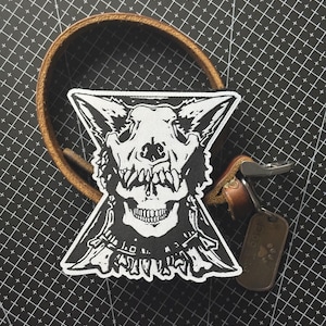 Pode incluir: Uma coleira de cão de couro castanho com um patch preto e branco com um crânio e características semelhantes às de um lobo. Uma etiqueta de metal com uma pegada de pata está pendurada na coleira.