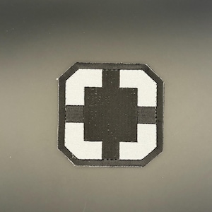 Könnte beinhalten: Quadratischer Aufnäher mit einem schwarzen Kreuzdesign auf weißem Grund. Der Aufnäher hat einen schwarzen Rand und ist vor einem dunkelgrauen Hintergrund platziert. Das Design ist eine einfache, geometrische Form.