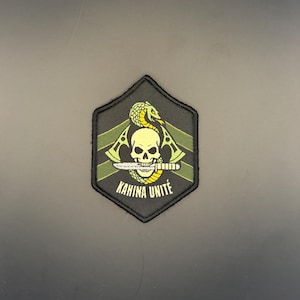 Op de afbeelding: Een zwarte patch met een gele schedel doorboord door een mes, met een geel-groene slang erboven. De tekst "KAHINA UNITÉ" staat in het wit. De patch heeft een zwarte rand en groene details.