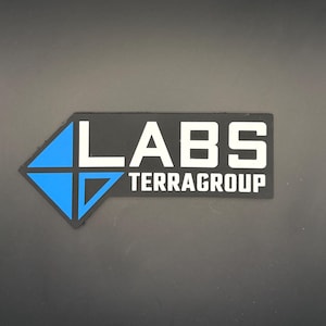 May include: A rectangular black patch featuring a blue diamond-shaped icon on the left side. The text LABS is printed in large white capital letters, with the word TERRAGROUP in smaller white capital letters positioned directly underneath it.
