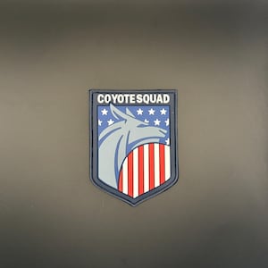 May include: A patch with a shield design featuring a gray wolf head, blue background with white stars, and red and white stripes. The text "COYOTE SQUAD" is at the top of the patch.