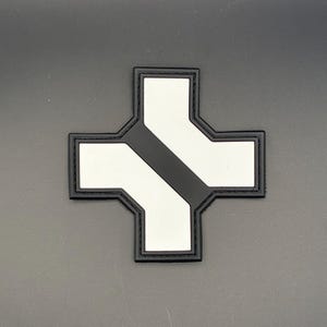 Könnte beinhalten: Ein schwarz-weißes Abzeichen in Form eines Kreuzes. Das Kreuz hat einen schwarzen Rand und einen schwarzen diagonalen Streifen in der Mitte. Der Hintergrund ist einfarbig grau.