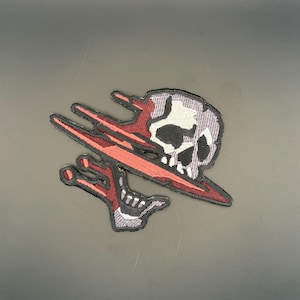 May include: Embroidered patch featuring a skull with a red slash across it. The skull is white and gray with black accents. The patch has a black border and red and black details.