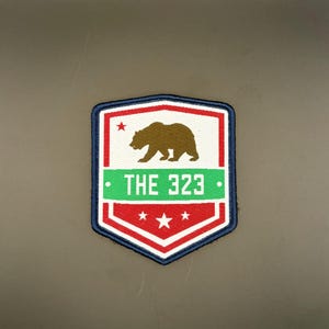 May include: A patch with a shield design featuring a brown bear silhouette, red and white accents, and the text "THE 323" in white on a green stripe. The patch has a blue border and red stars.