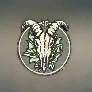 May include: A circular embroidered patch featuring a white goat skull with black outlines centered over light green foliage. The design is set against a dark gray circular background with a thin light green border, creating a detailed graphic emblem.