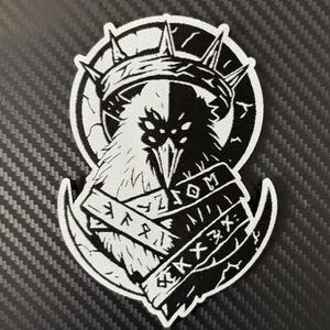 May include: A black and white patch featuring a raven with a crown, split vertically. The raven is set against a circular background. Below the raven are two banners with stylized text. The patch has a white border.