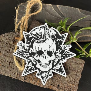 Può includere: Una toppa in bianco e nero con un teschio e decorazioni floreali, incorniciata da una forma a stella. La toppa è presentata su un pezzo di legno strutturato, con un anello di spago attaccato.