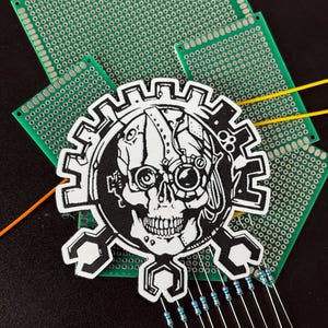 Könnte beinhalten: Ein schwarz-weißer Aufnäher mit einem Schädel mit mechanischen Details, eingebettet in ein Zahnraddesign. Der Aufnäher liegt auf einer grünen Leiterplatte, mit Widerständen und Drähten. Dunkler Hintergrund.