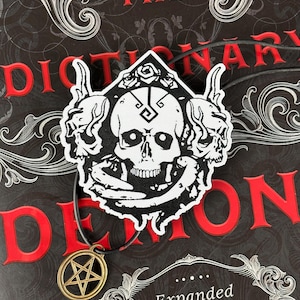 Könnte beinhalten: Eine Halskette mit Anhänger in Schwarz und Weiß, die einen Totenkopf mit Hörnern und einen Pentagramm-Anhänger zeigt. Der Anhänger befindet sich an einer schwarzen Kordel. Der Hintergrund ist ein Buchcover mit "THE DICTIONARY OF DEMONS" in Rot.