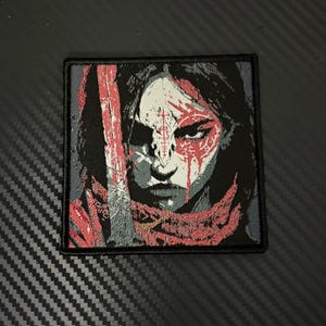 May include: Square patch featuring a stylized portrait of a warrior with red paint markings on the face and a red scarf. The warrior has a stern expression, with black hair and a gray background.