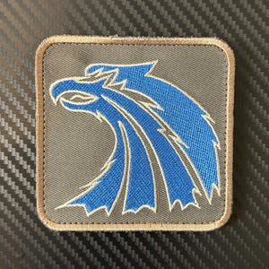 Pode incluir: Patch quadrado com uma cabeça de águia azul estilizada sobre um fundo cinza. O desenho da águia é delineado em branco e o patch tem uma borda bege. Ideal para roupas ou acessórios.