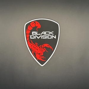 Può includere: Una toppa a forma di scudo con sfondo nero e bordo bianco. Un grafico di uno scorpione rosso brillante è posizionato sul lato sinistro. Il testo BLACK DIVISION è stampato in lettere bianche, maiuscole e senza grazie al centro.