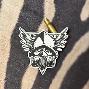 May include: A black and white sticker featuring a gas mask-wearing figure with wings. The sticker has a triangular shape and is set against a zebra-striped background. A gold bullet casing is also present.