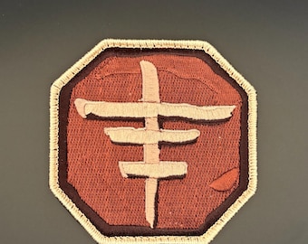May include: Embroidered patch with a geometric design in shades of brown and beige. The patch has a dark brown border and a beige stitched edge. The design is centered on a reddish-brown background.