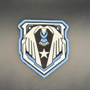 Könnte beinhalten: Ein schildförmiger, bestickter Aufnäher mit einem stilisierten weißen Adler mit ausgebreiteten Flügeln auf schwarzem Grund. Das Design umfasst einen zentralen weißen Stern, zwei kleinere Sterne oben und einen hellblauen Rand.