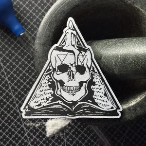 Op de afbeelding: Een zwart-wit patch met een schedel in een driehoek, met een kaars en geometrische vormen. Het ontwerp bevat een open boek en is waarschijnlijk voor kleding of accessoires.