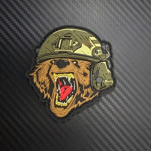 May include: A patch featuring a roaring bear wearing a military helmet and headset. The bear has brown fur, a wide open mouth with a red tongue, and sharp white teeth. The helmet and headset are in shades of green and tan.