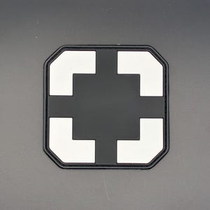 May include: A black and white patch featuring a cross design. The patch has a black background with a white cross shape in the centre. The patch is rectangular with rounded corners.