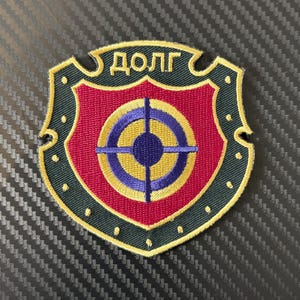 Pode incluir: Patch bordado em forma de escudo com um centro vermelho e um desenho de alvo em azul e amarelo. O escudo é contornado em verde e dourado, com a palavra cirílica "ДОЛГ" no topo.
