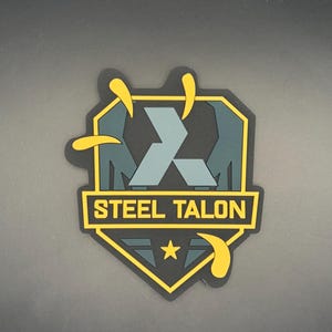 Könnte beinhalten: Ein schildförmiger Aufnäher mit schwarzem Hintergrund und einem dunkelblauen geometrischen Design. Der Text STEEL TALON ist in gelben Blockbuchstaben auf einem horizontalen Balken gedruckt. Drei gelbe Krallenspuren und ein kleiner Stern.