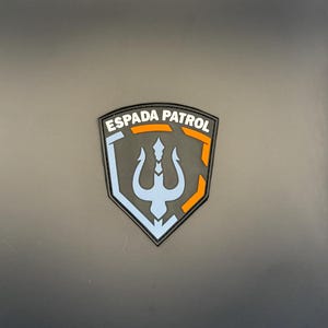 Może przedstawiać: Czarna i szara naszywka z napisem "ESPADA PATROL" w kolorze białym i pomarańczowym. Naszywka przedstawia symbol trójzębu w kolorze jasnoniebieskim i wzór tarczy z pomarańczowymi akcentami.