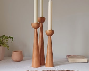 May include: Three wooden candle holders with a tapered design and a rounded top. The candle holders are made of natural wood and have a warm, rustic look. The candle holders are arranged in a row on a white tablecloth.