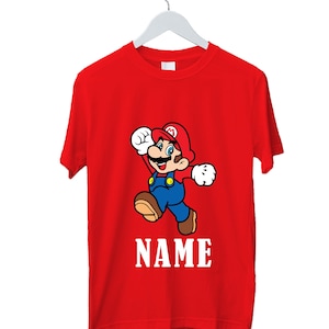 May include: Red t-shirt with a cartoon illustration of Mario, wearing a red hat and blue overalls, in a running pose. The word "NAME" is printed in white below the character. The t-shirt is displayed on a white hanger.