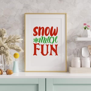 May include: A framed print with the words "Snow Much Fun" in red and green. The print is in a gold frame and is displayed on a white countertop. The background is a white tile wall. Decorative items are on the countertop.