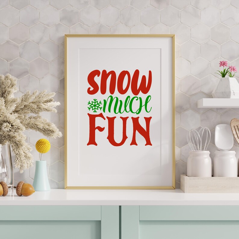 May include: A framed print with the words "Snow Much Fun" in red and green. The print is in a gold frame and is displayed on a white countertop. The background is a white tile wall. Decorative items are on the countertop.