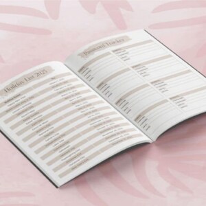 May include: Open notebook with a black cover, displaying a "Holiday List 2025" and "Password Tracker" on its pages. The pages have a beige and white color scheme with text. The notebook is on a pink surface.