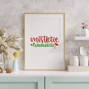 May include: A framed print with the words "mistletoe & margaritas" in red and green. The text is in a decorative font, with a cocktail glass icon. The frame is gold, and the background is white. The print is displayed on a light blue surface.