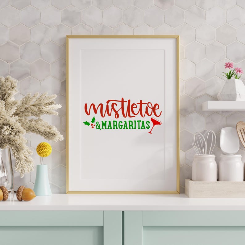 May include: A framed print with the words "mistletoe & margaritas" in red and green. The text is in a decorative font, with a cocktail glass icon. The frame is gold, and the background is white. The print is displayed on a light blue surface.