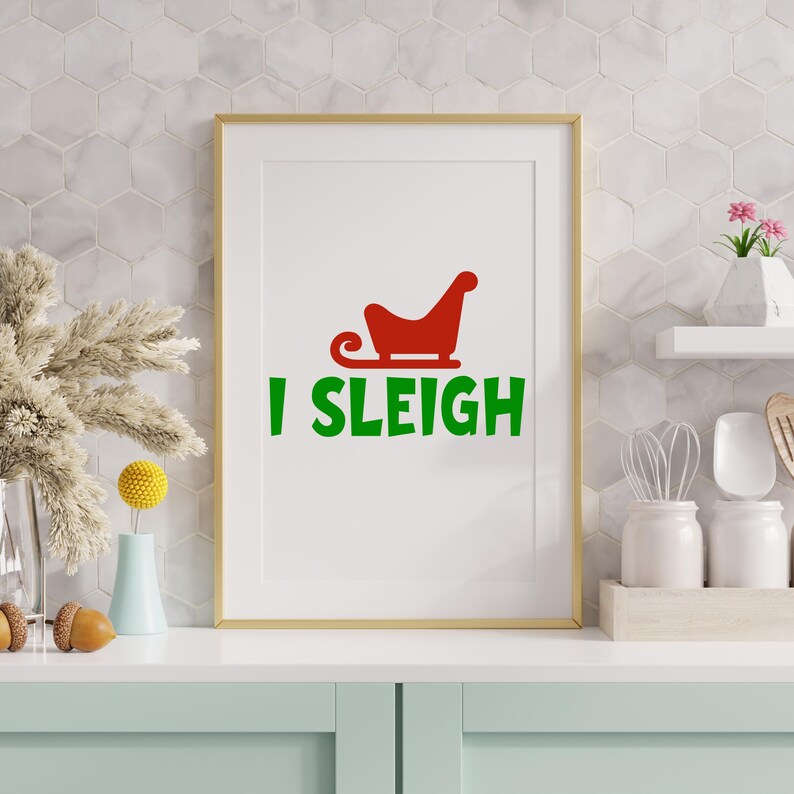 May include: A framed print with a gold border features a red sleigh graphic above the green text "I SLEIGH" on a white background. The print is displayed on a light blue surface with decorative items.