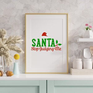May include: A framed print with a gold border displaying the text "SANTA Stop Judging Me" in green and red. The design includes a Santa hat and holly accents, ideal for festive decorations. The print is displayed on a white surface.