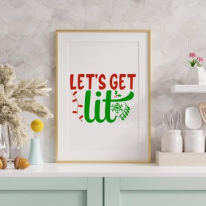 May include: A framed print with the text "LET'S GET lit" in red and green. The word "lit" is stylized with Christmas lights and a snowflake design. The frame is gold, and the print is displayed on a white surface.