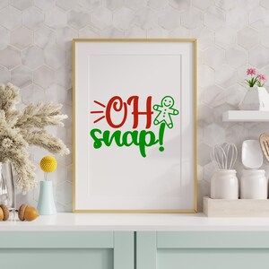 May include: A framed print with the words "OH snap!" in red and green, and a gingerbread man graphic. The print is in a gold frame. The background features a white tile wall. The print is displayed on a white surface.