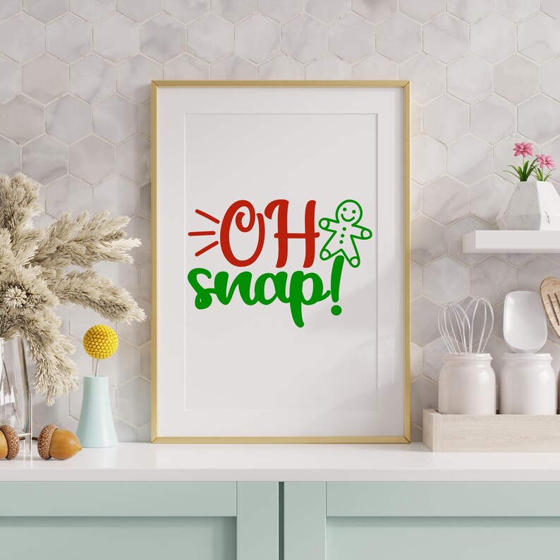 May include: A framed print with the words "OH snap!" in red and green, and a gingerbread man graphic. The print is in a gold frame. The background features a white tile wall. The print is displayed on a white surface.