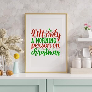 May include: A framed print with the text "I'm only a morning person on Christmas" in red and green. The print is in a gold frame and is displayed on a white surface with decorative items.