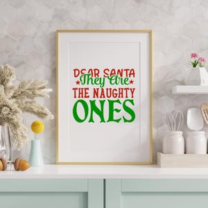 May include: A framed print with the text "DEAR SANTA They Are THE NAUGHTY ONES" in red and green. The framed art is in a gold frame and is displayed on a white countertop with decorative items.