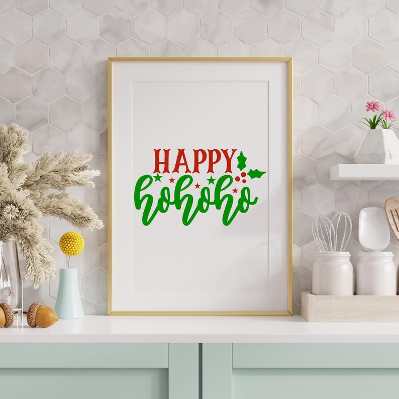 May include: A framed print with the words "HAPPY hohoho" in red and green. The gold-framed artwork is displayed on a white surface with decorative items, including a vase with dried plants and a small vase with a yellow flower.