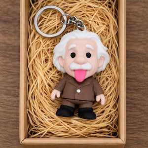 Può includere: Un portachiavi a forma di cartone animato di Albert Einstein con la lingua di fuori. La figura ha i capelli bianchi, un abito marrone e scarpe nere. È presentato in una piccola scatola di cartone piena di paglia.