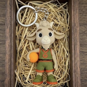 Könnte beinhalten: Ein Schlüsselanhänger mit einem ziegenartigen Charakter, der einen orangefarbenen Basketball hält, in einer Holzkiste mit Stroh. Der Charakter trägt eine grüne und orangefarbene Basketballuniform. Der Schlüsselanhänger hat einen silbernen Ring.
