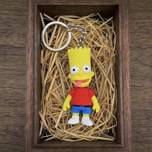Może przedstawiać: Brelok do kluczy Bart Simpson ze srebrnym kółkiem i łańcuszkiem. Figurka jest żółta z czerwoną koszulką, niebieskimi szortami i białymi butami. Brelok jest prezentowany w drewnianym pudełku wypełnionym słomą.