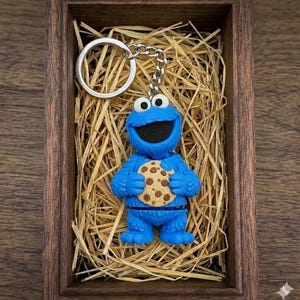 Puede incluir: Un llavero azul del Monstruo de las Galletas sosteniendo una galleta con chispas de chocolate. El llavero está hecho de un material azul con detalles en blanco y negro. El llavero se exhibe en una caja de madera con paja.